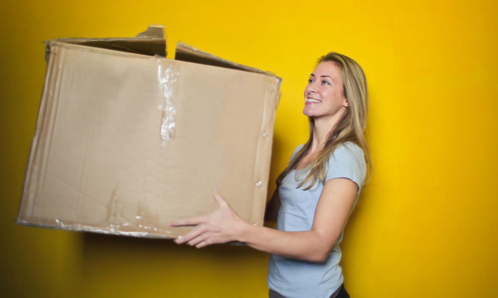 Your Renters Insurance Survival Guide - Woman Carrying Very Large Box Against a Yellow Wall